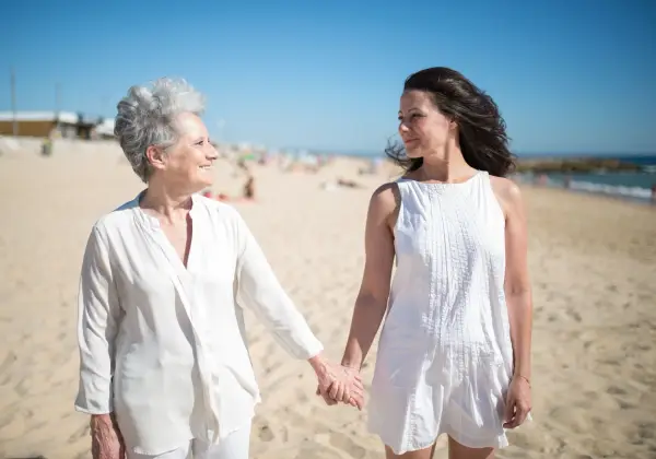 An adult daughter and her elderly mother holding hands on the beach