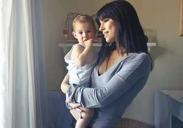 A mother holding her toddler by a sunlit window