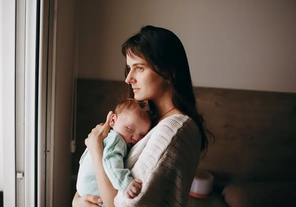 A weary mother holding her sleeping newborn by a window