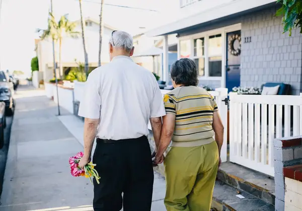 An older husband and wife walking together, he carrying flowers