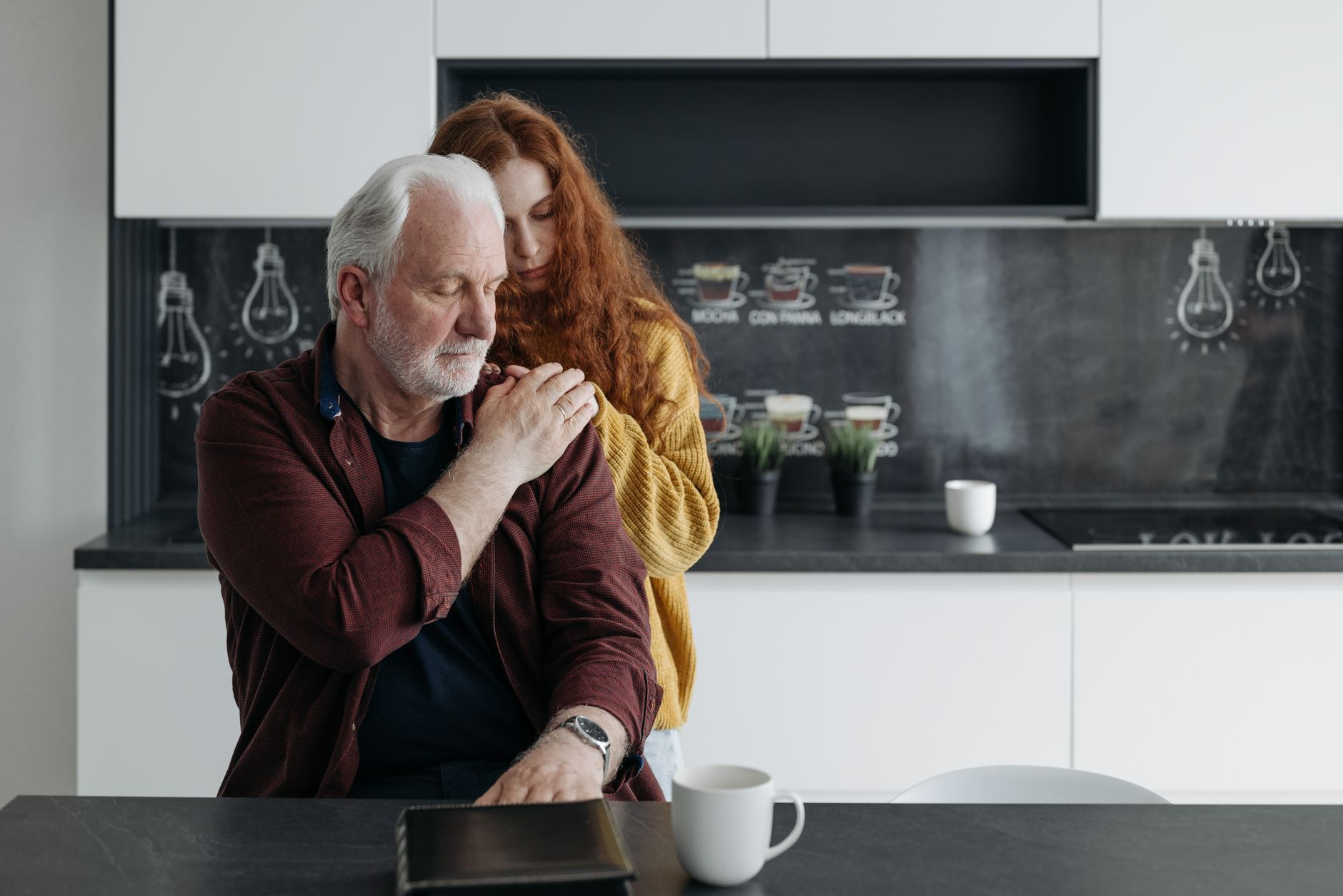 A daughter embracing her aging father from behind in the kitchen
