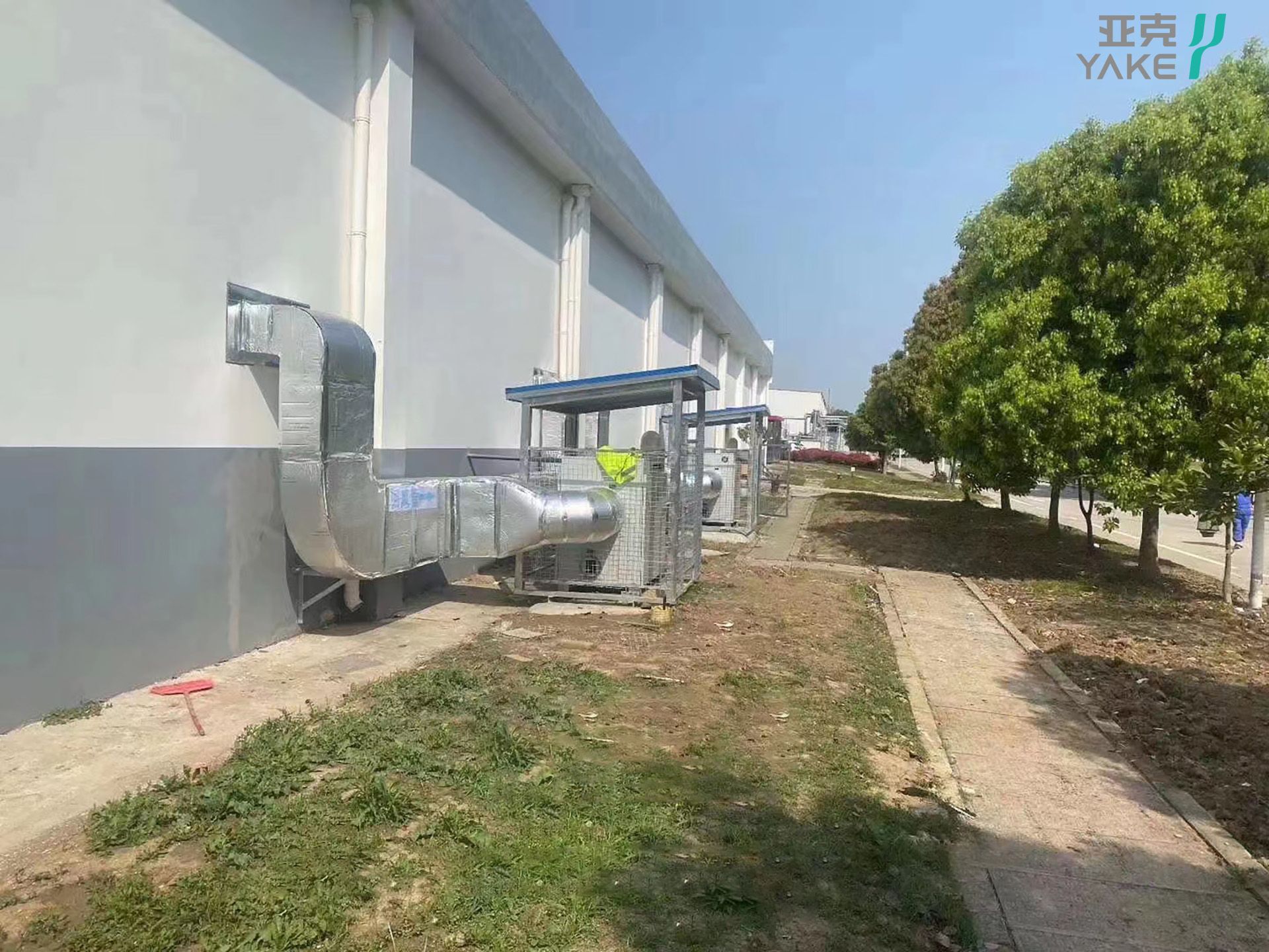 A building exterior with large metal ventilation ducts, likely part of a Yakeclimate Industrial Dehumidifier system, extends from the wall. A worker in a safety vest appears to be inspecting the dehumidification installation. This setup reflects advanced dehumidification manufacturing by a renowned Commercial Dehumidifier Manufacturer. The area is surrounded by grass and trees under a clear blue sky, highlighting the sustainable setting of this industrial site. Yakeclimate Dehumidification Manufacturing