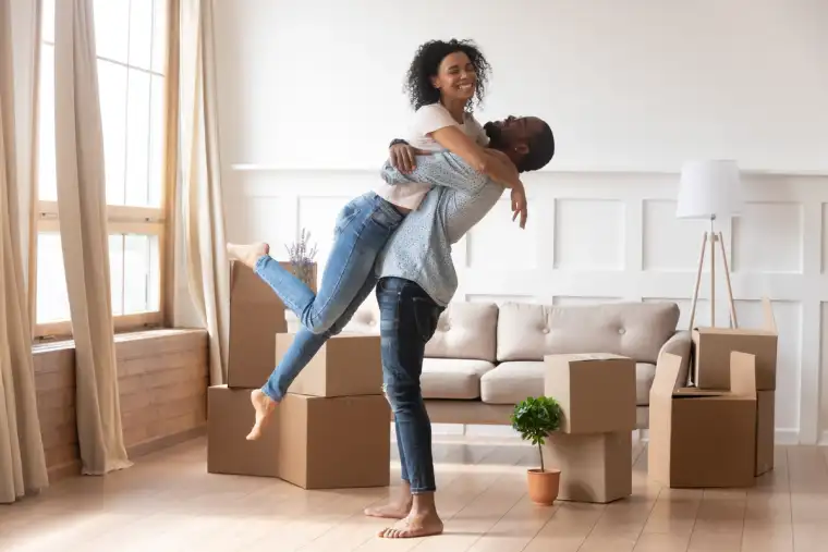 First time home buyers celebrate new house purchase on moving day, smiling husband embracing lifting excited young wife laughing standing among boxes in own house