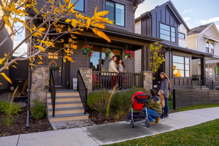 Family chatting with neighbours on a front porch in an Alpine Park laned home, with a stroller on the sidewalk and fall trees lining the street