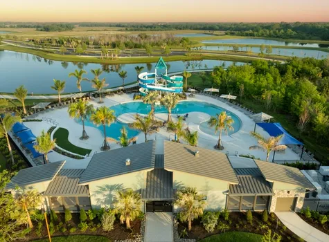Aerial view of Camp Creek at North River Ranch featuring a resort-style pool with palm trees, cabanas, and a twisting water slide, surrounded by scenic lakes and lush greenery at sunset.