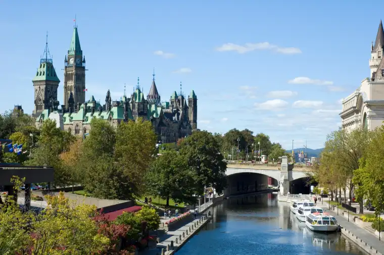 Ottawa skyline featuring Parliament Hill with its iconic Peace Tower, overlooking the Rideau Canal with boats, a bridge, and tree-lined pathways under a clear blue sky.