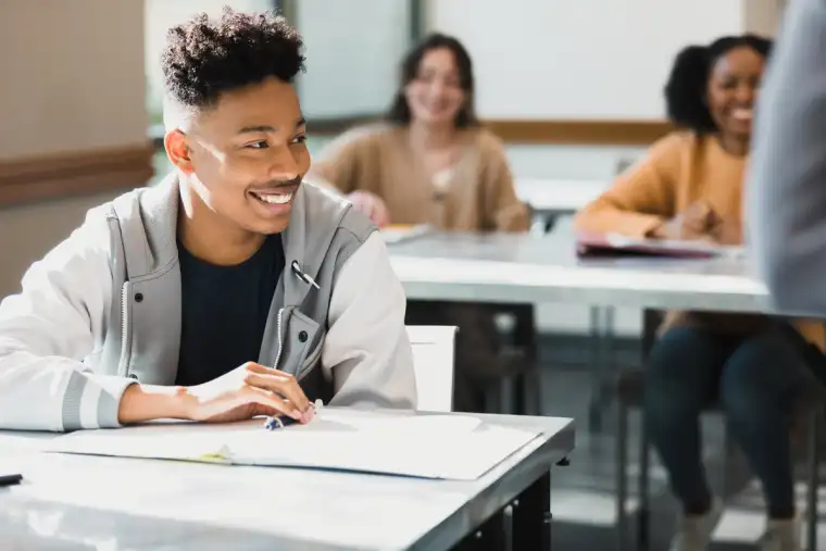 Student smiling and listening in a bright classroom, with classmates in the background taking notes and engaging in a lesson