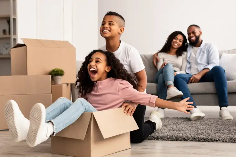 Happy family having fun and celebrating moving day, excited brother and sister playing with cardboard box, parents hugging while sitting on the couch in background