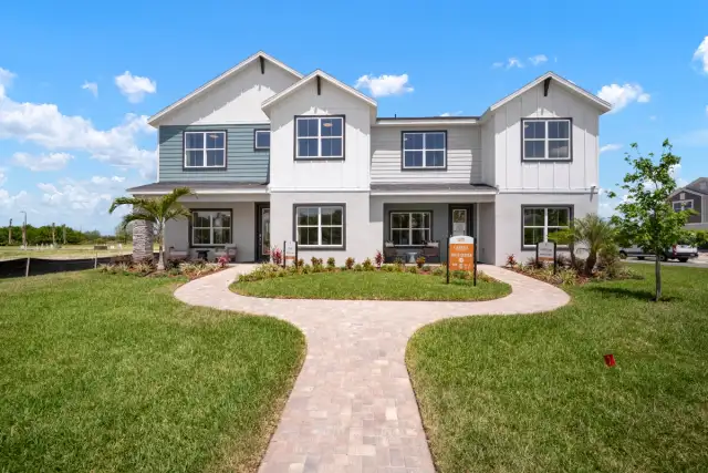 Modern two-storey paired model home exterior with white board-and-batten siding, black-framed windows, landscaped front entry, curved stone walkway, and manicured green lawn under a bright blue sky.