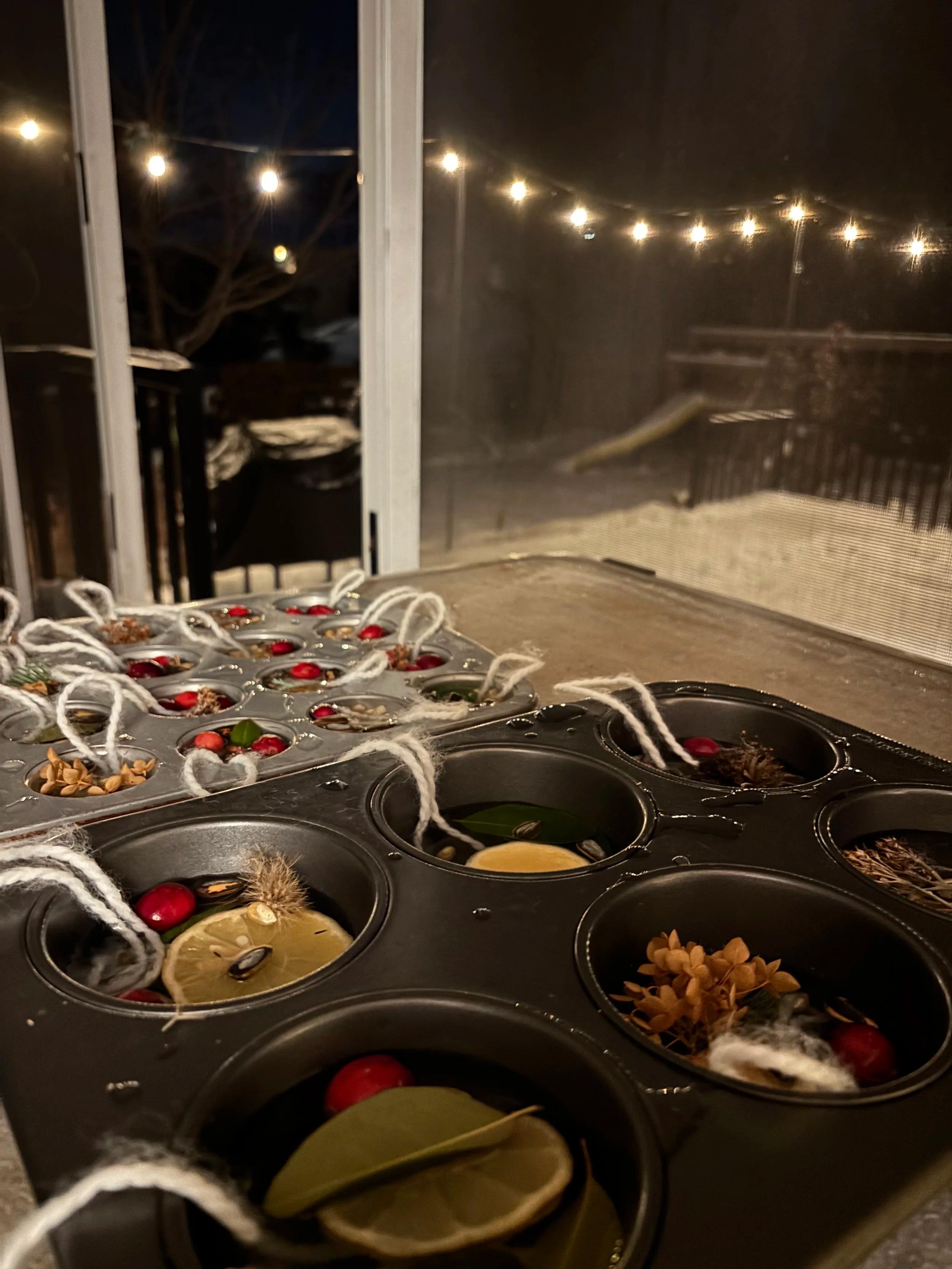 Two muffin tins full of ornament supplies set out on a wintry Minnesota deck with lights, snow, and a swing set in the background.