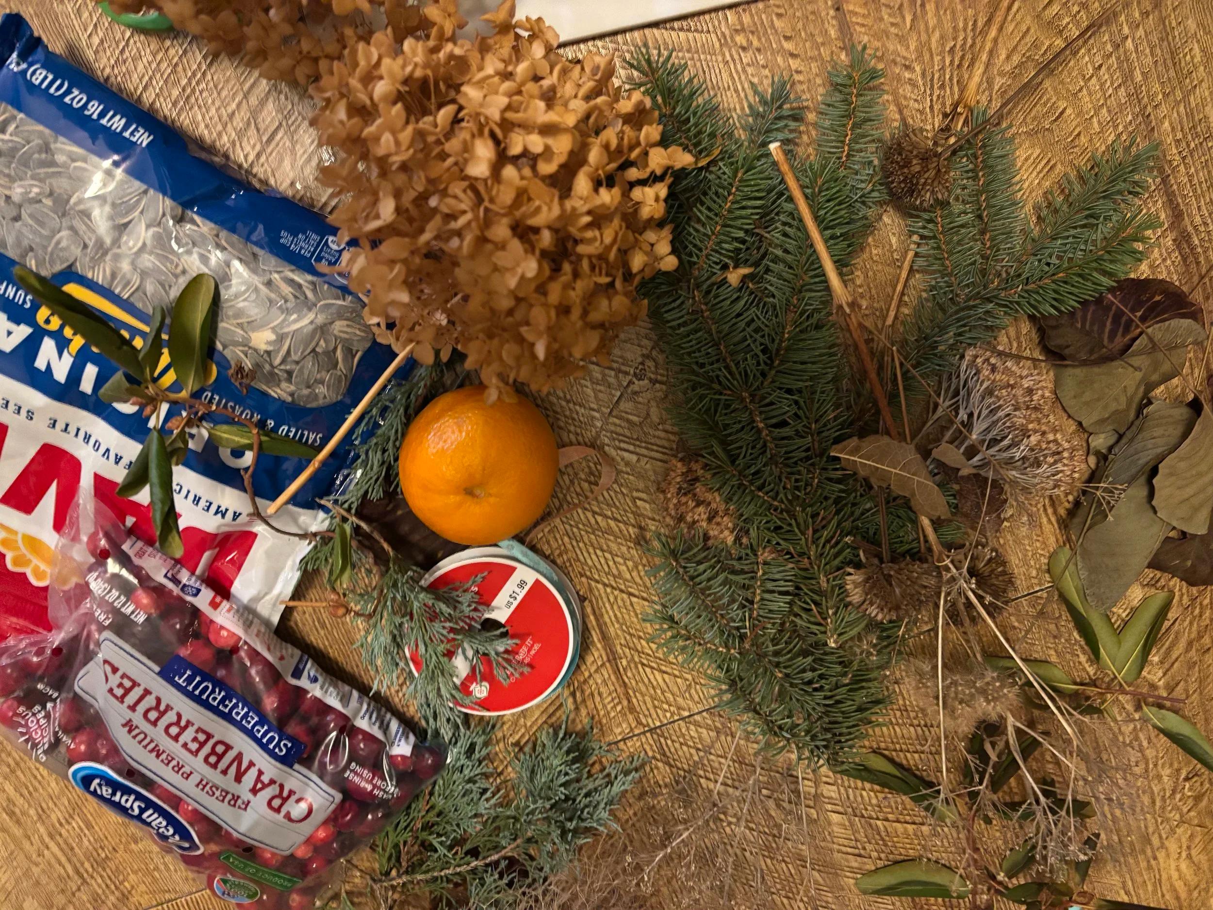 pine boughs, leaves, an orange, cranberries, and sunflower seeds on a table with ribbon.