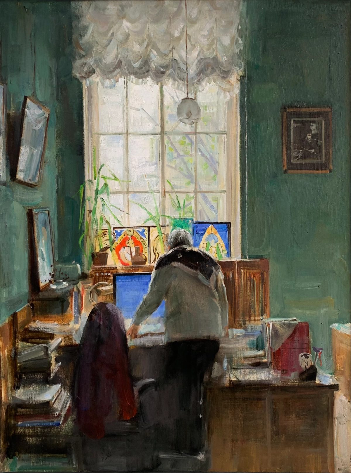 Man standing at a desk in an office with a lace-curtained window and paintings on the sill.