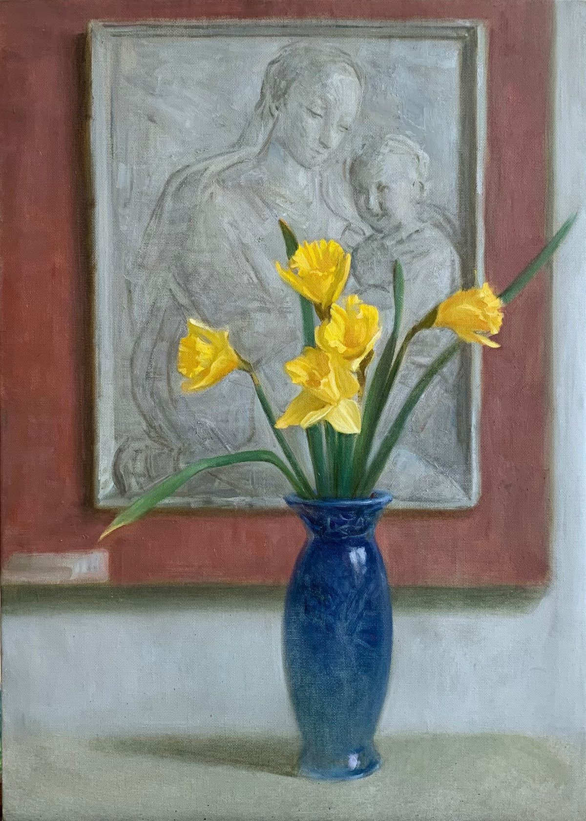 Yellow daffodils in a blue vase before a framed portrait on a red wall.