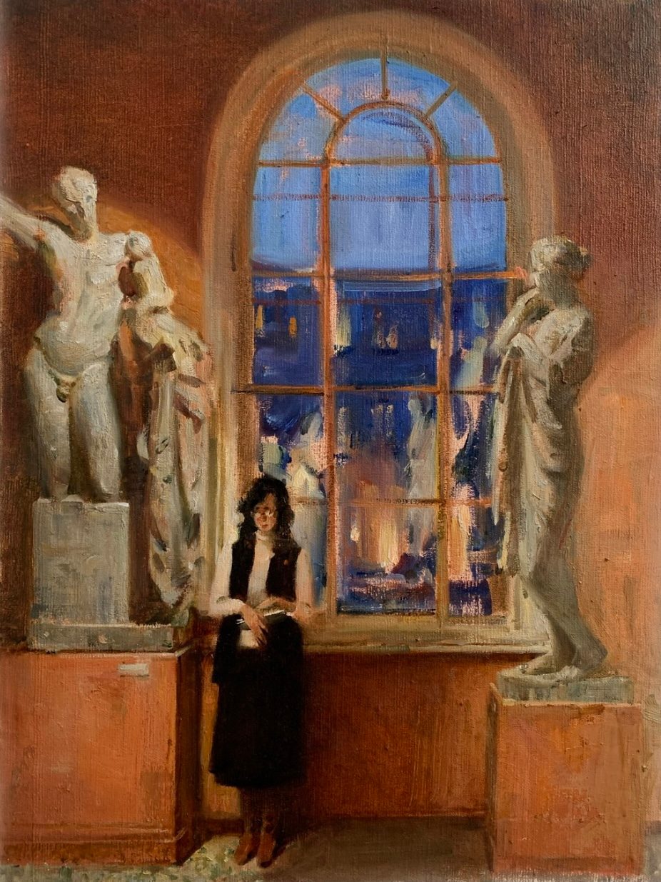 Woman in a black dress seated between plaster sculptures before a tall arched window at dusk.