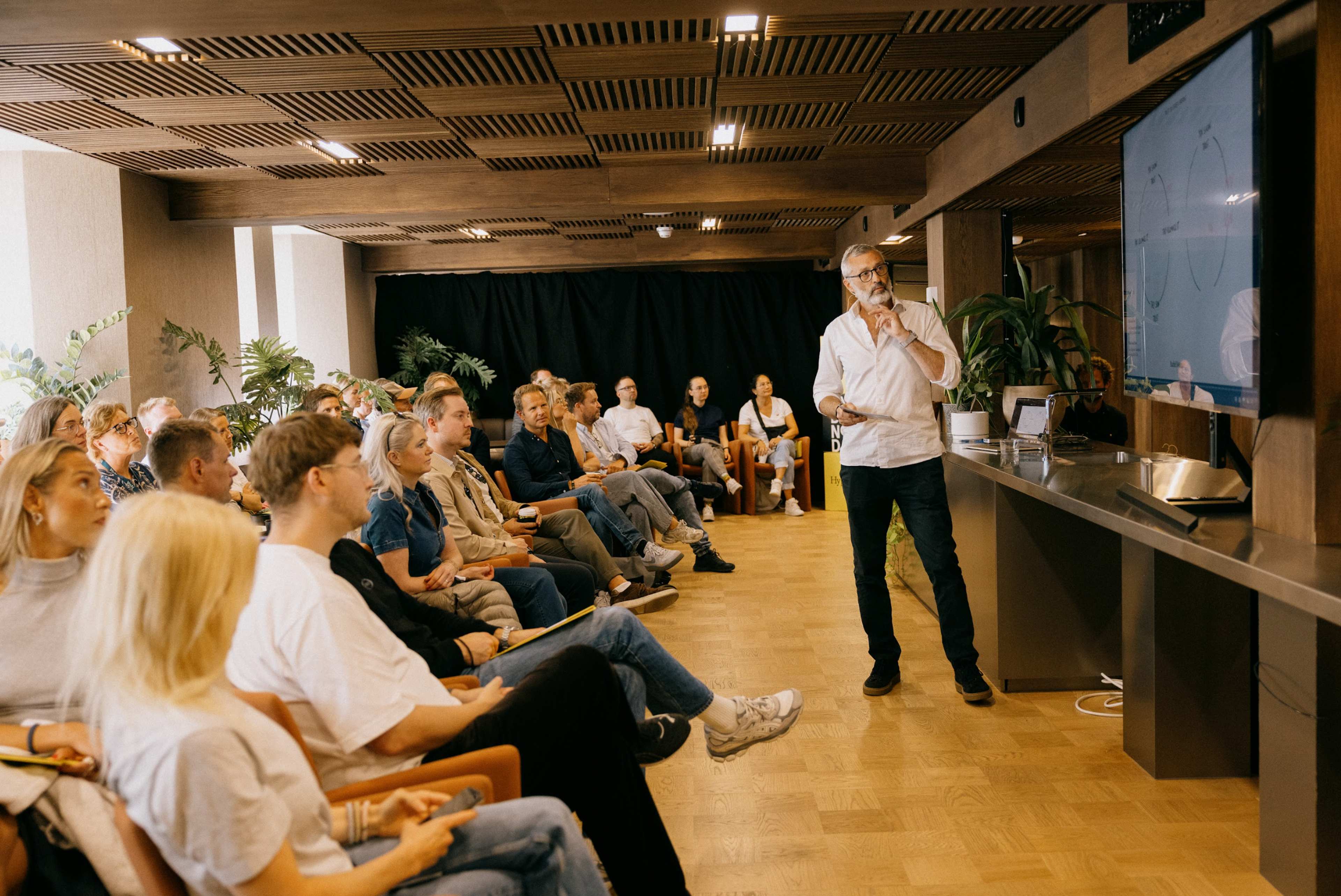 A man presents to an audience in a room with a large screen.