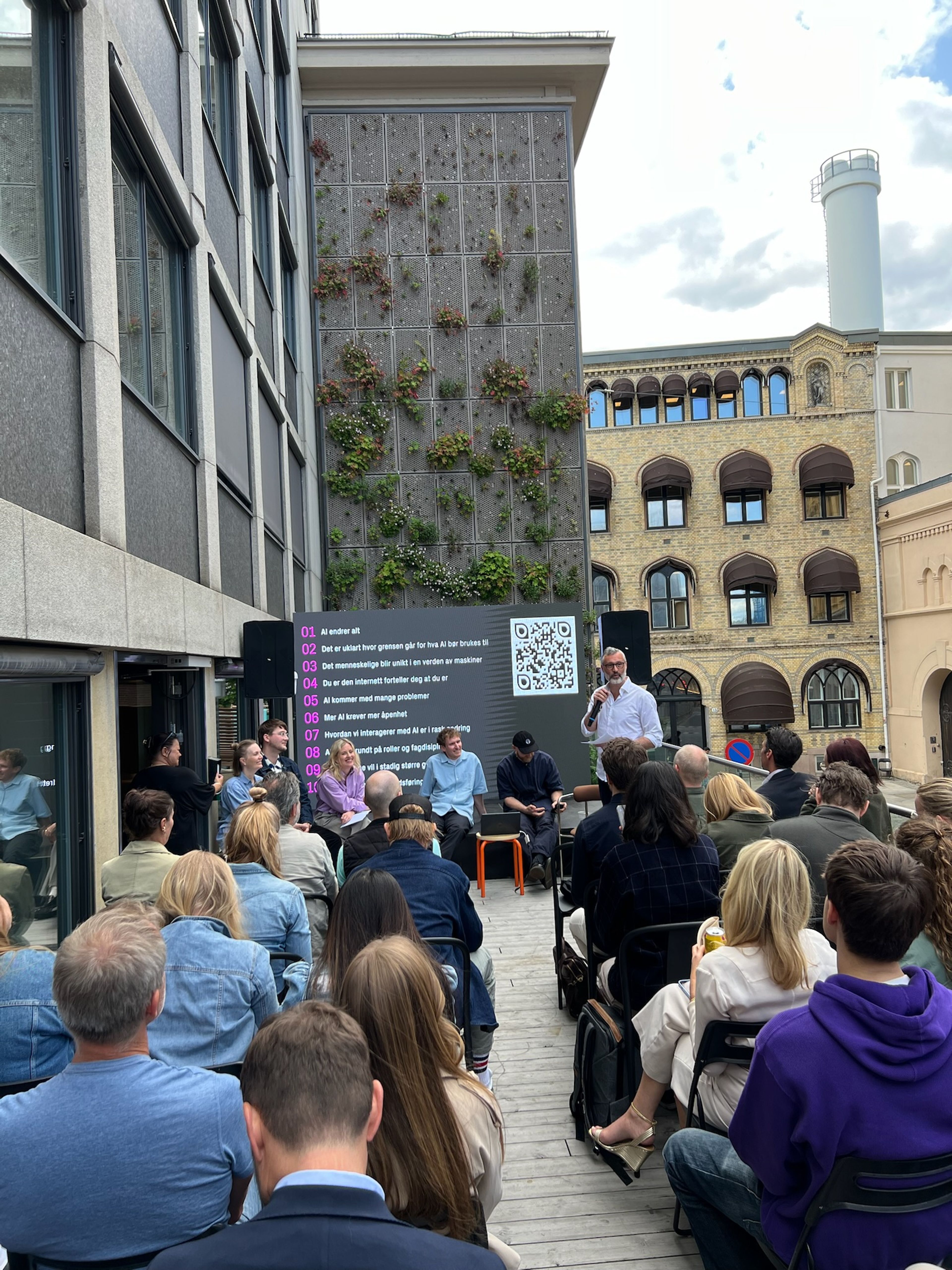 A speaker addresses a seated audience and a panel at an outdoor event, with a building featuring a large green living wall behind them.