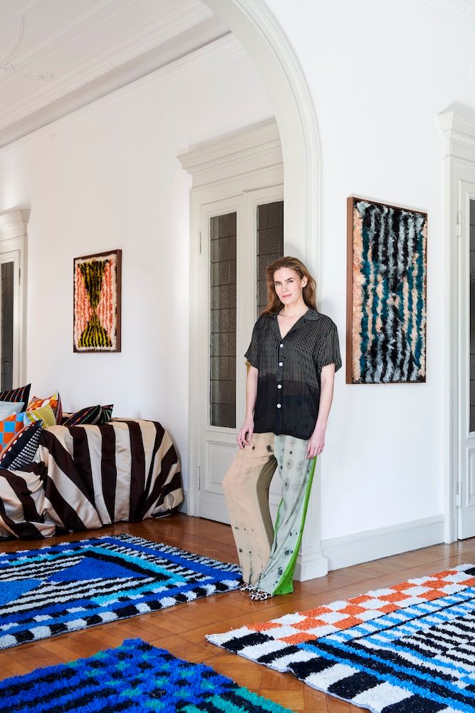 A woman in a black sheer shirt and mismatched pants stands in a room with colorful patterned rugs, a striped sofa, and abstract wall art.