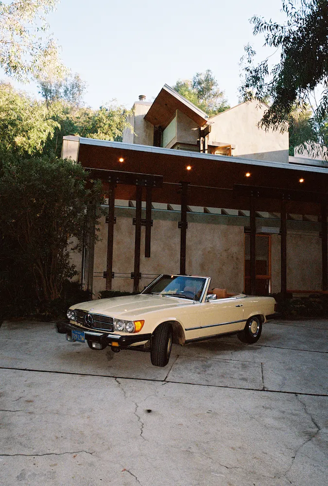 A classic car in cream in front of a house with concrete floor in the foreground
