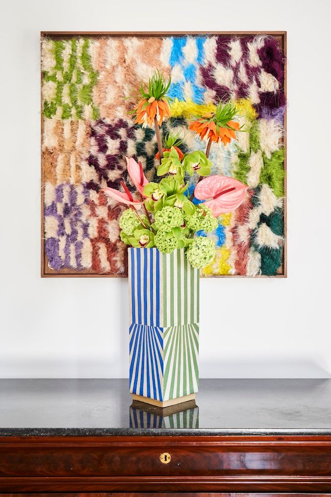 A tall, striped blue and green vase filled with orange, green, and pink flowers sits on a dark table in front of a framed, multicolored, textured patchwork wall art.
