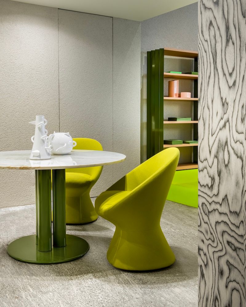 A modern interior with a white marble table on a green base, two bright yellow chairs, a tall green bookshelf, and a black and white patterned wood panel.