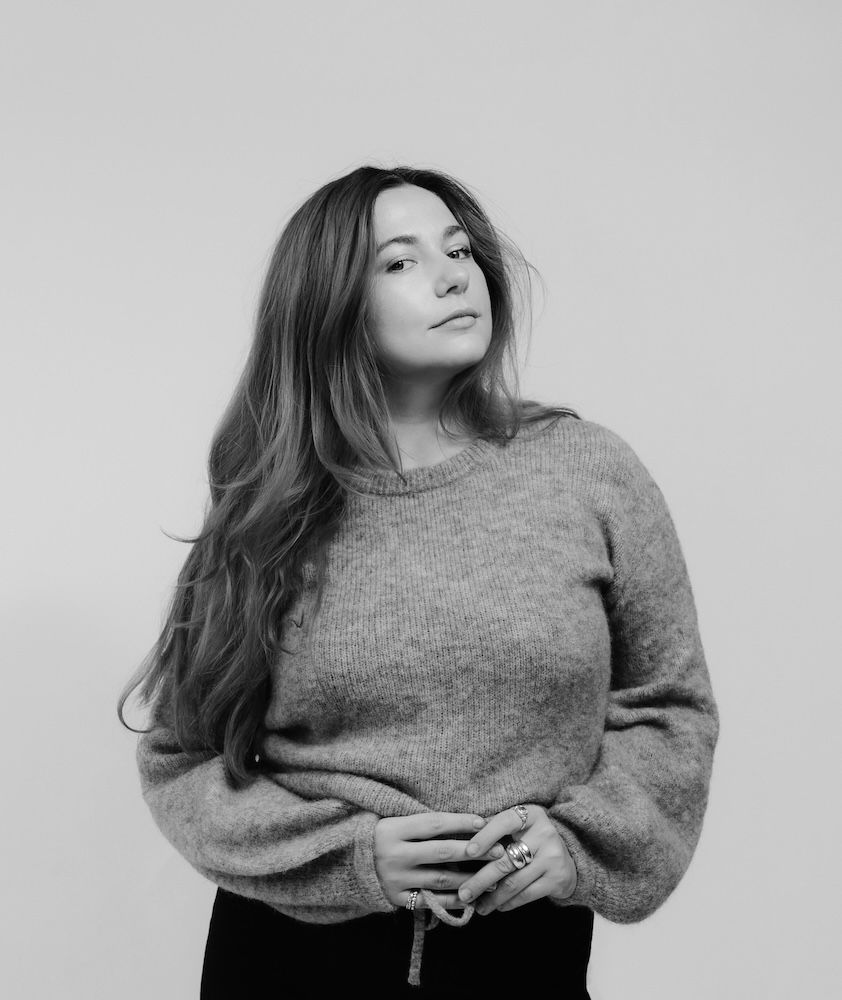 Monochrome portrait of a woman with long, wavy hair wearing a fuzzy sweater, looking at the camera with her hands clasped.