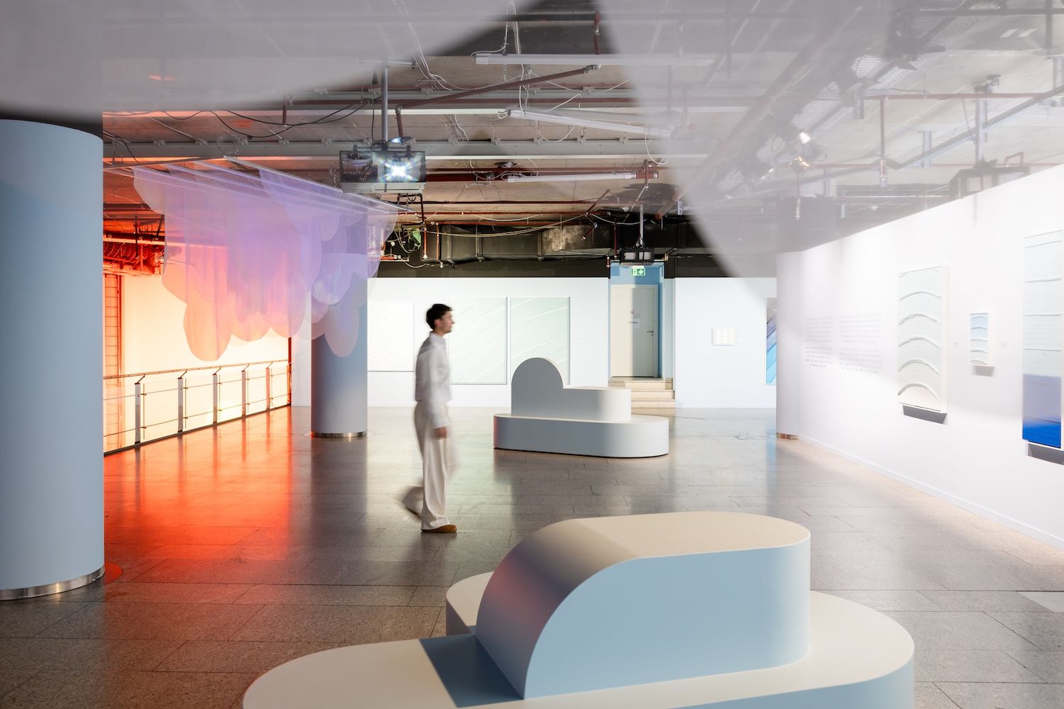 Man walking through exhibition set up with seating and artworks on wall in muted pastel colours and brightness on the left