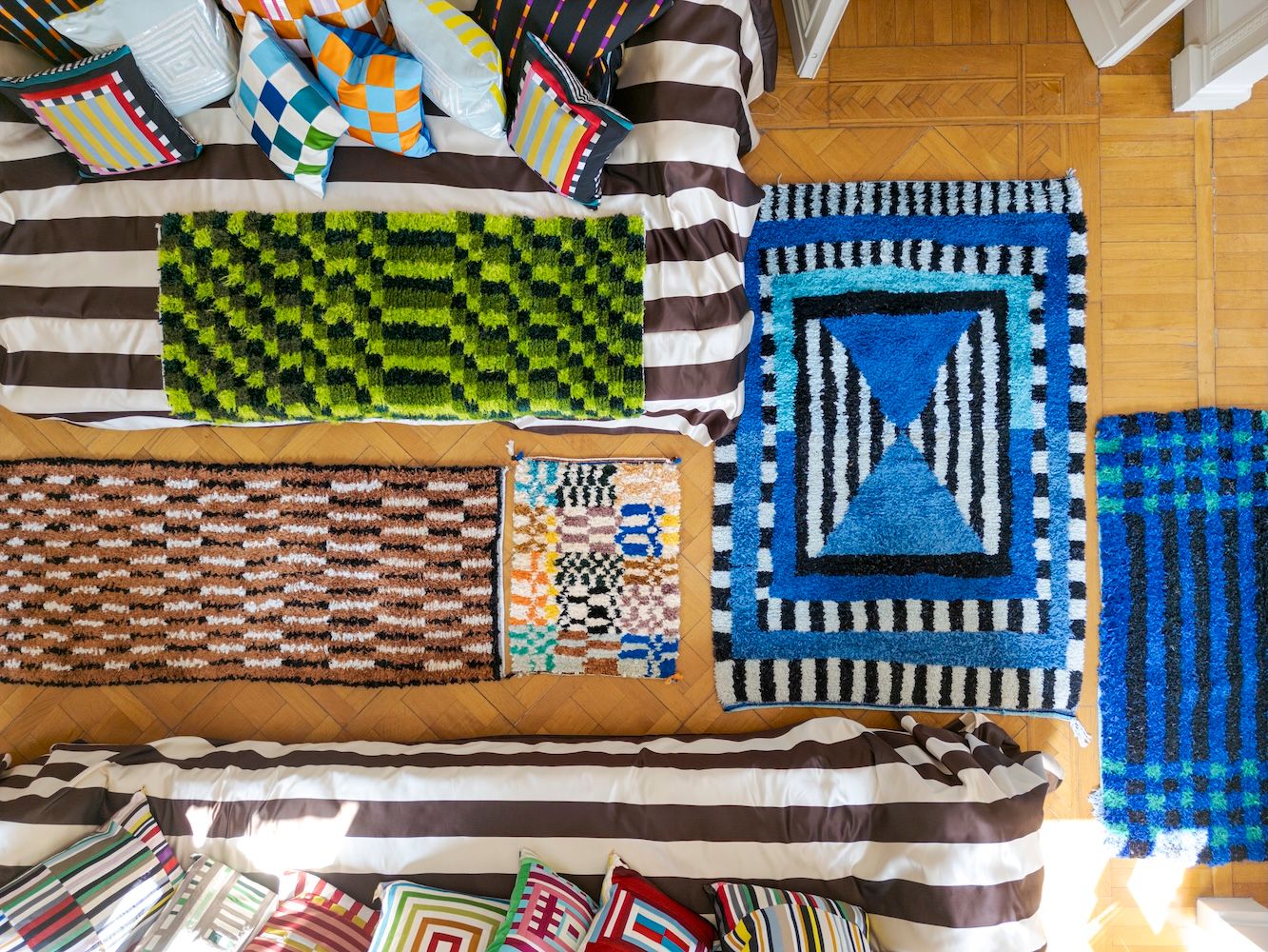 Overhead view of several vibrant, textured rugs with geometric patterns displayed on a wooden floor, alongside a striped bed with colorful pillows.