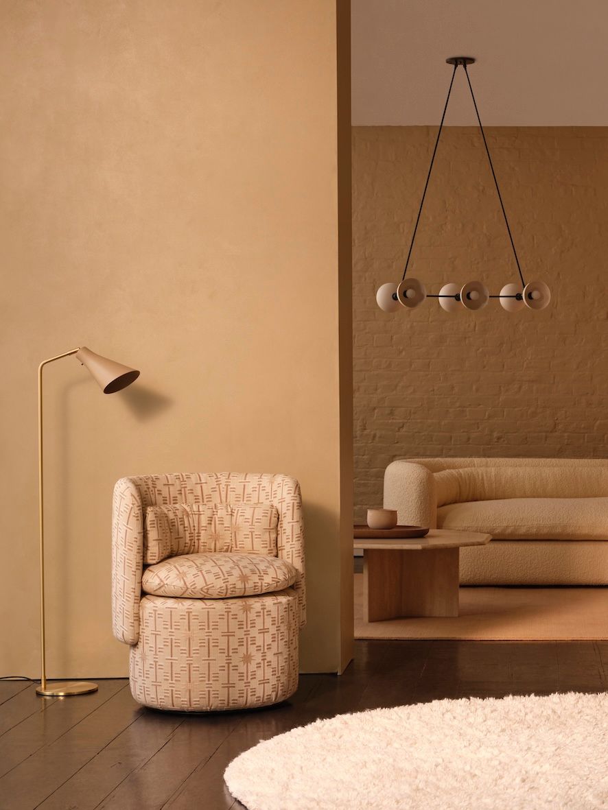 A warm, minimalist living space featuring a patterned swivel chair, a gold floor lamp, a neutral-toned sofa, and a modern pendant light.