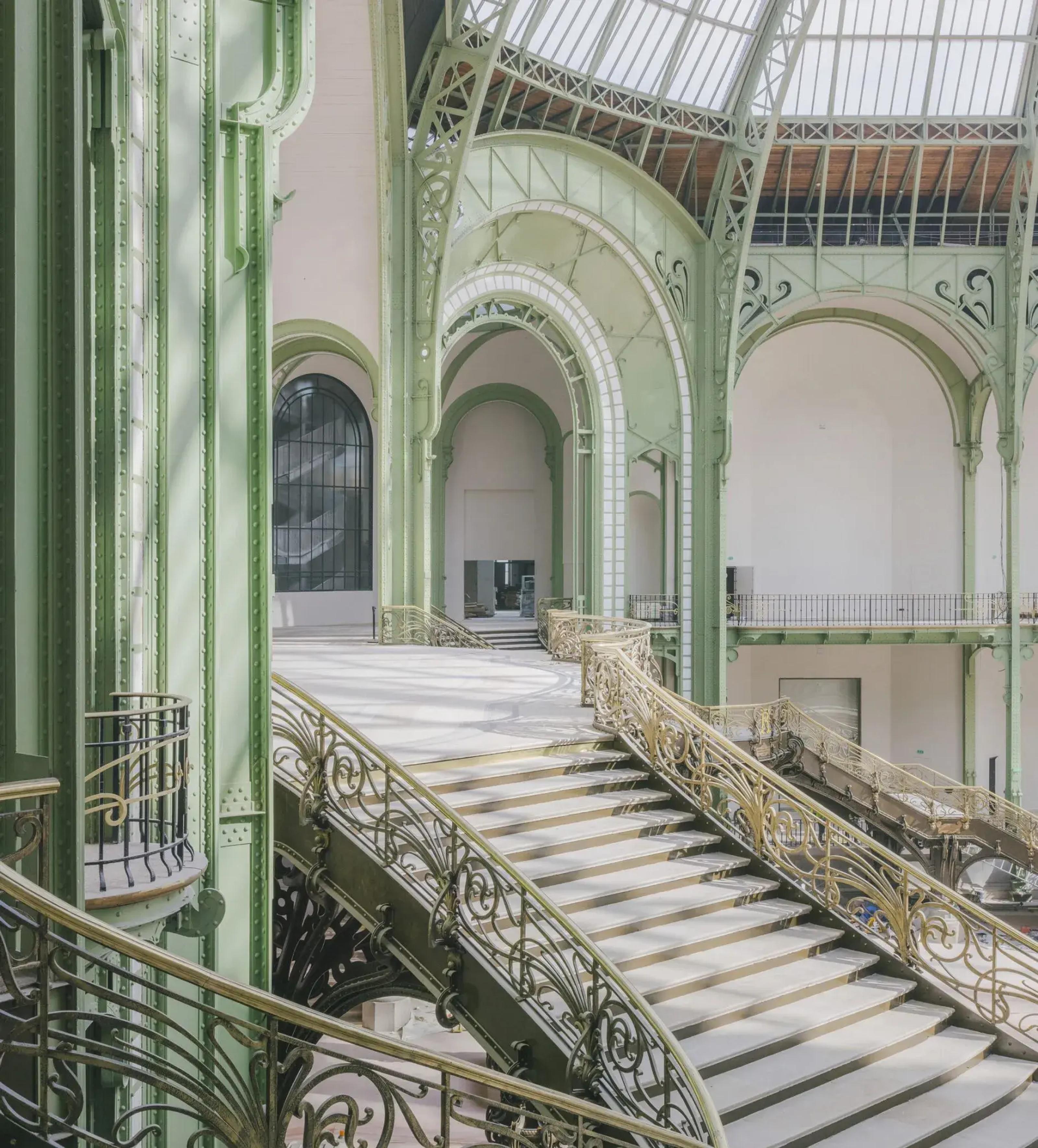 Elegant staircase in the Grand Palais with intricate wrought iron railings, soft green arches, and natural light streaming through a glass dome