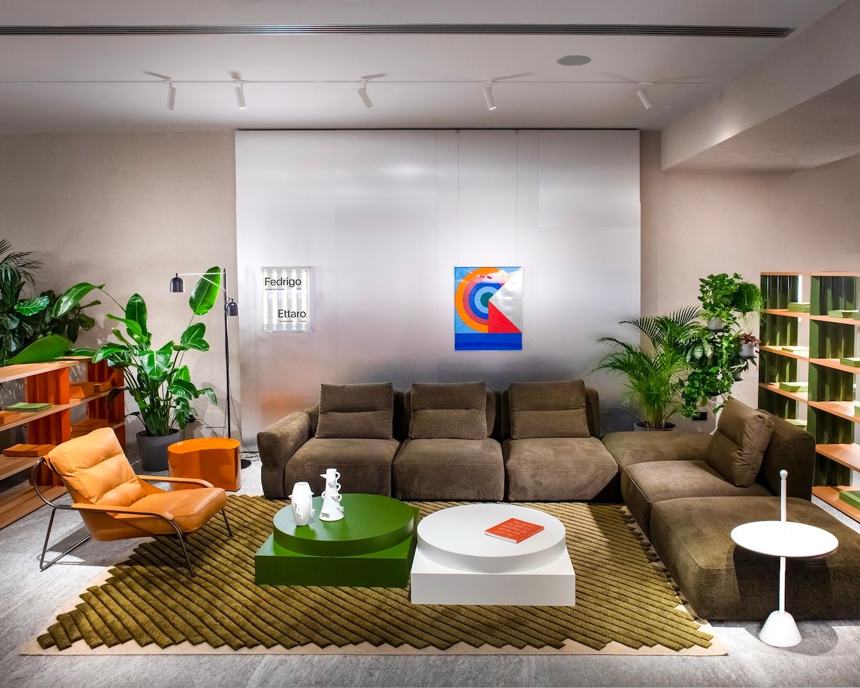 A modern interior featuring a large brown sectional sofa, an orange armchair, green and white coffee tables, a green geometric rug, and potted plants.