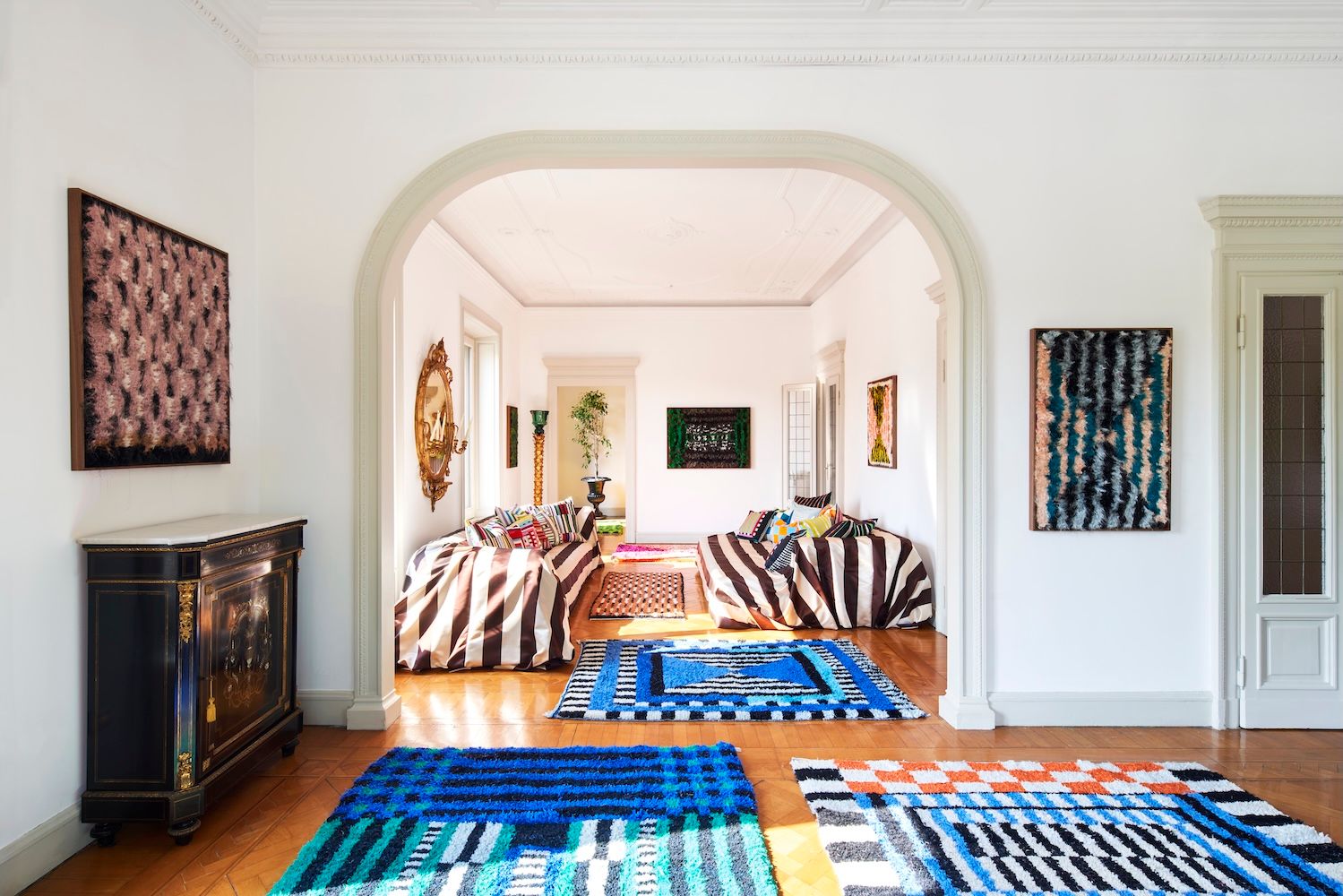 A brightly lit room with white walls, wooden floors, colorful geometric rugs, striped sofas, and abstract art.