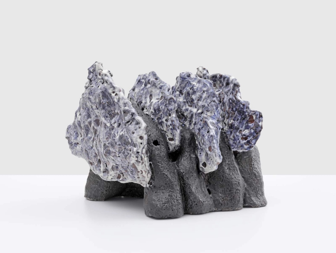 A sculpture with a base resembling gray rock formations topped with textured, white and blue elements, displayed on a white surface against a plain background.