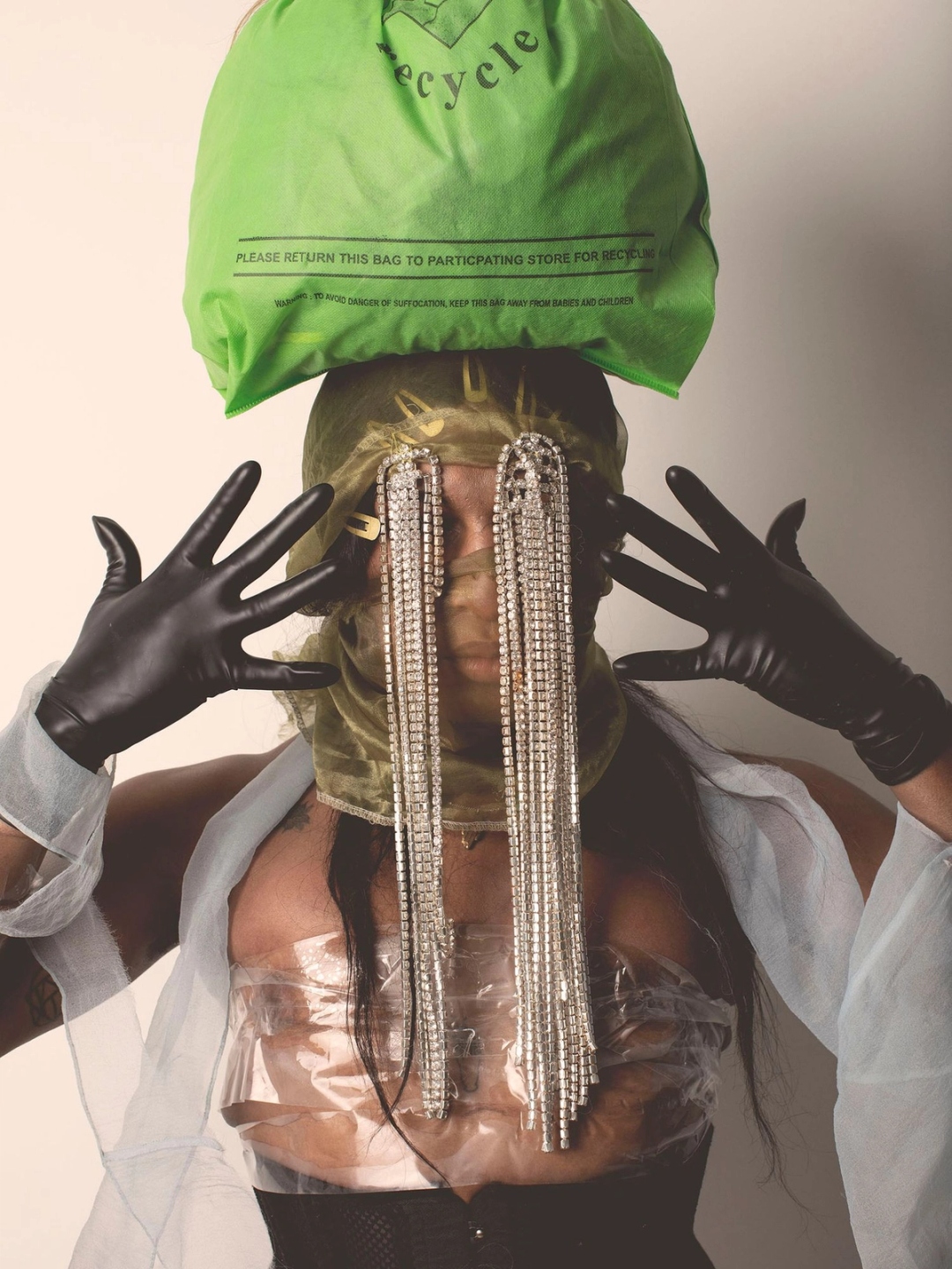 A photograph of a figure posing with their hands framing their face with black gloves on. The figure had a green recycling bag atop their head, with a tulle head covering with long strings of gem attached to the head covering cascading down their face obscuring their face.