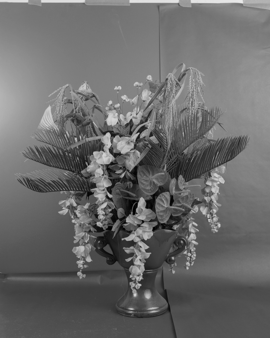 A black-and-white photo of a bouquet of artificial flowers, including bent wheat stalks cascading orchids, and anthurium lilies.