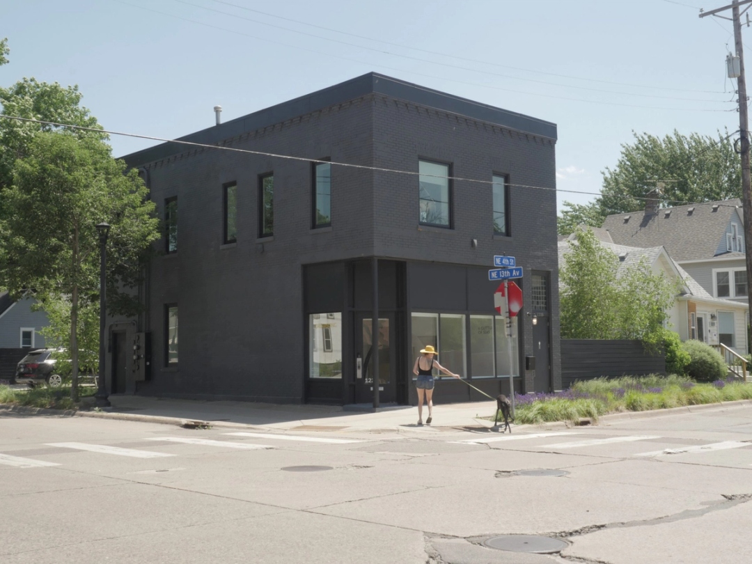 A woman wearing a sunhat walks past a modern, black-painted two-story art gallery on a sunny day, with a clear blue sky and suburban houses in the background.