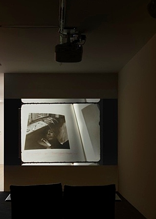 A still from Forks & Spoons showing a person below a windowsill, projected onto a wall. Two chairs are positioned in front of the projection.