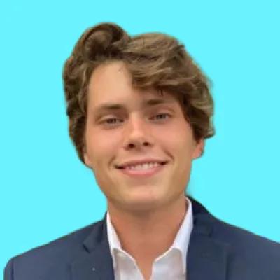A young man with light brown hair smiles, wearing a dark suit jacket and white shirt against a light blue background.