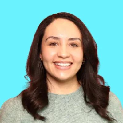 Headshot of a smiling woman with dark, wavy hair and a light green sweater against a light blue background.