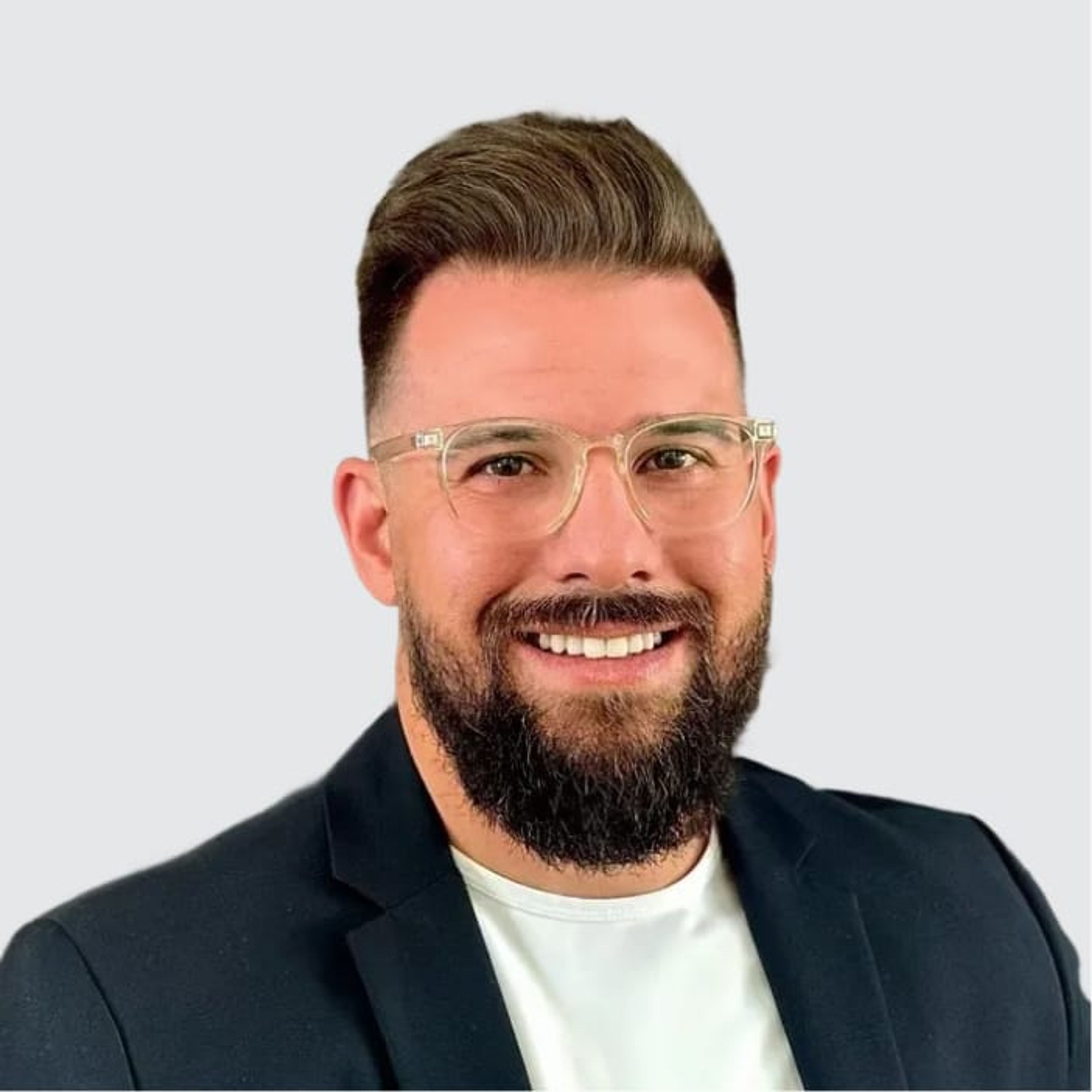 A smiling man with a beard and clear-frame glasses, wearing a black blazer over a white shirt.