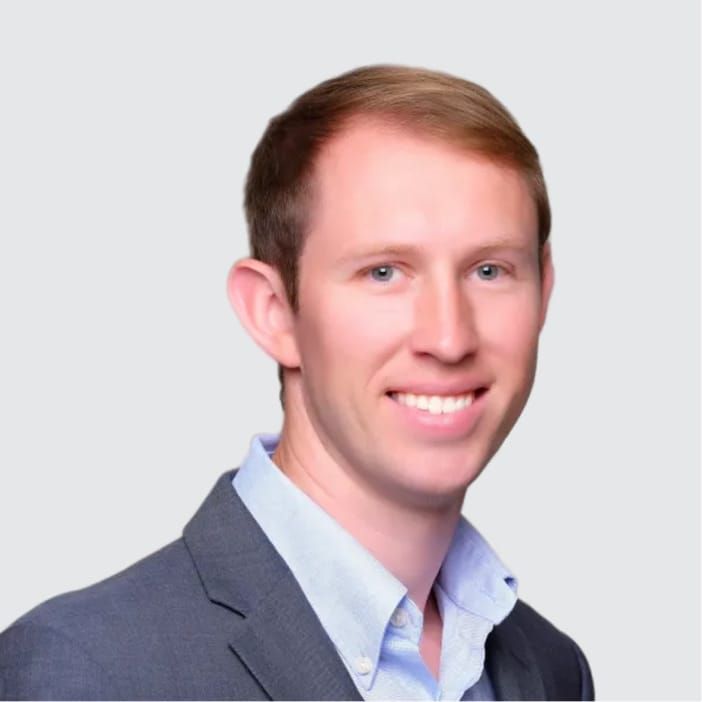 Smiling man with light brown hair and blue eyes, wearing a grey suit jacket and blue shirt.