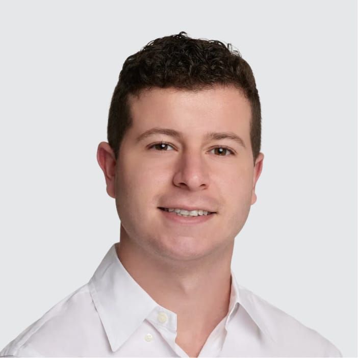 Headshot of a smiling young man with dark curly hair and a white shirt against a light gray background.