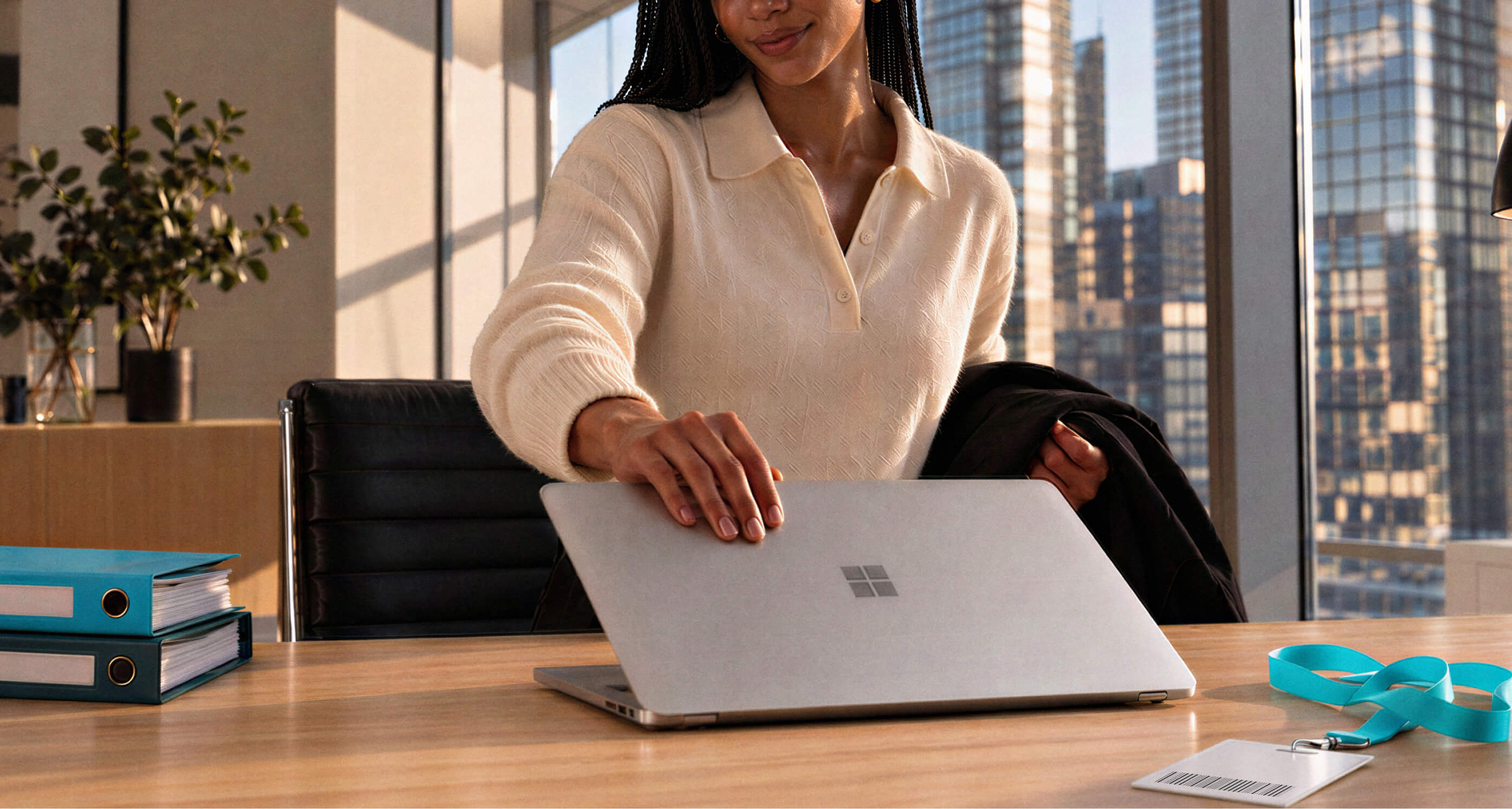 A woman opens a silver Microsoft Surface laptop on a wooden desk in an office with a cityscape view. Binders and an ID badge are visible.