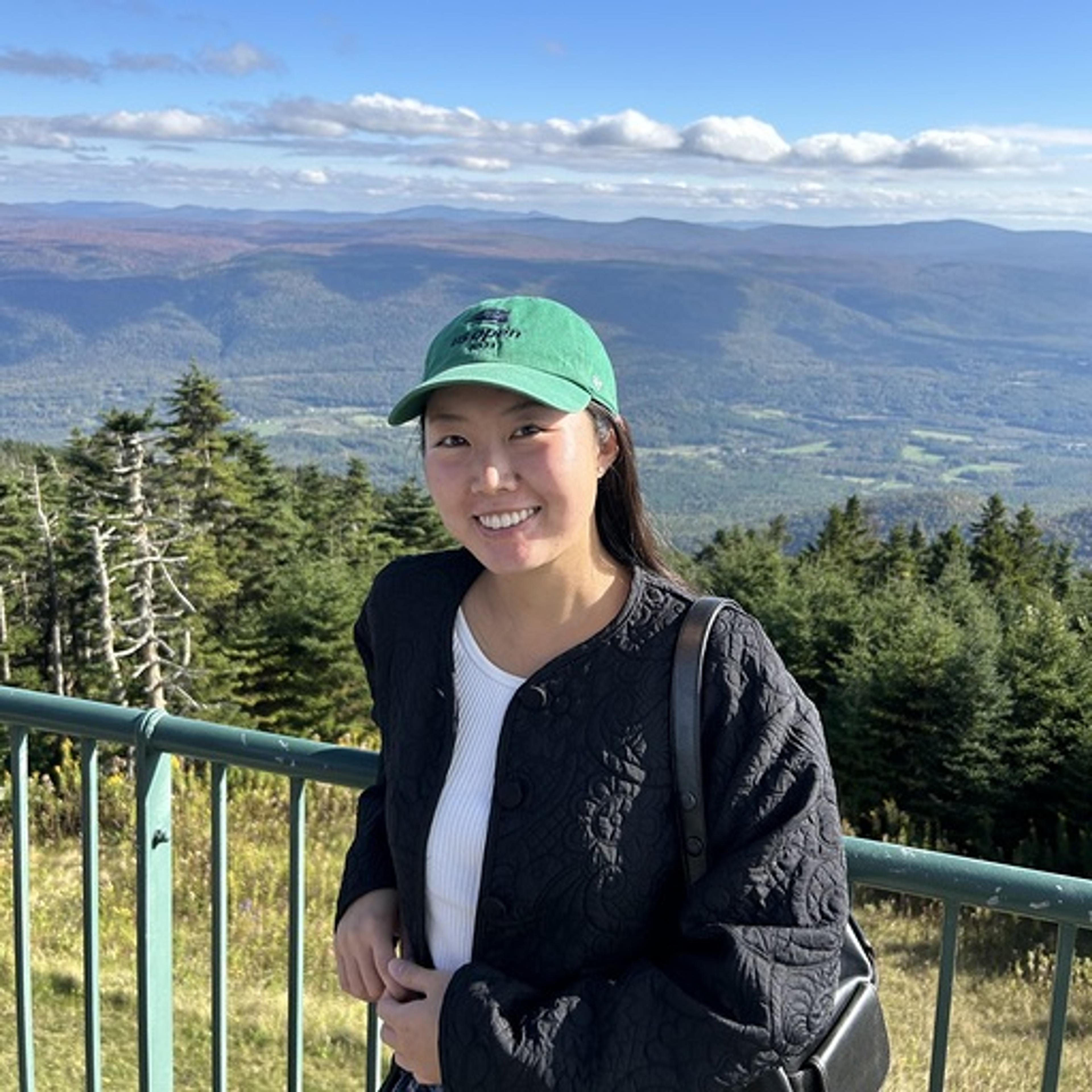 A smiling young woman in a green cap stands on a mountain overlooking a forested valley.