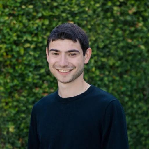 A young man smiling in a dark sweater against a green hedge.