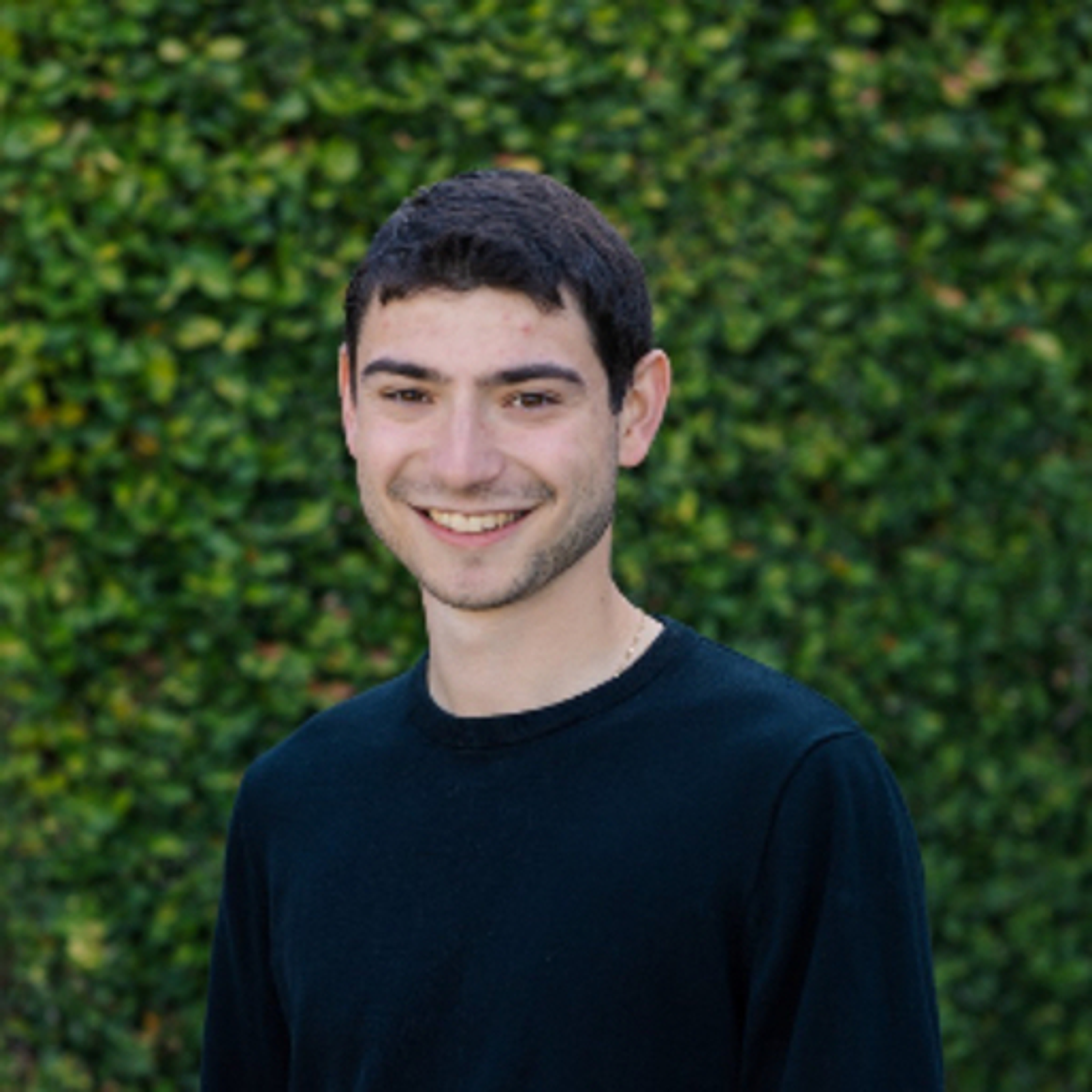 A young man smiling in a dark sweater against a green hedge.