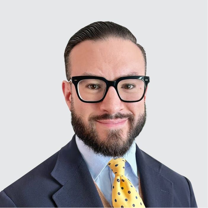 A smiling man with a beard and black glasses wearing a suit and yellow polka-dot tie.