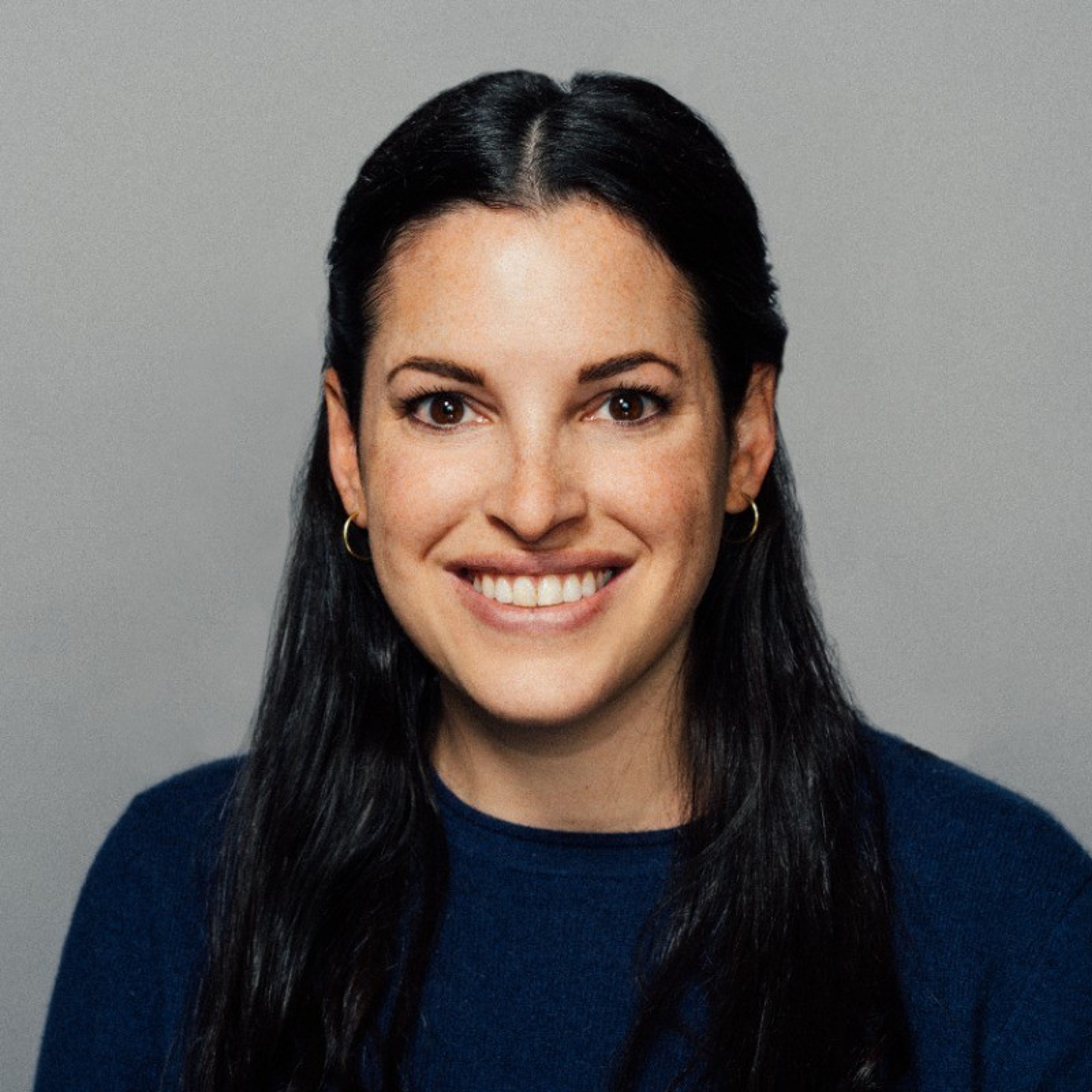 A smiling woman with long dark hair wears a navy blue top against a gray background.