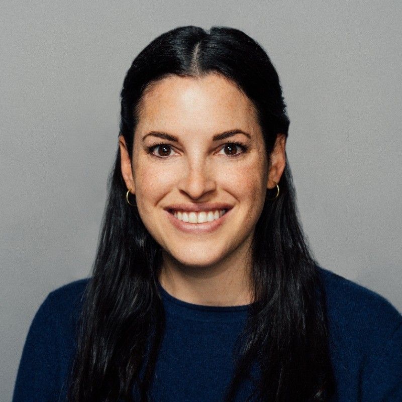 A smiling woman with long dark hair wears a navy blue top against a gray background.