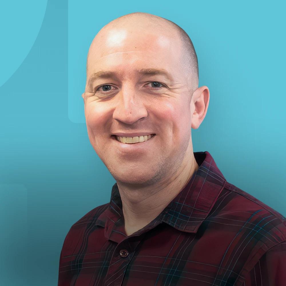 Smiling bald man in a red plaid shirt against a teal background.