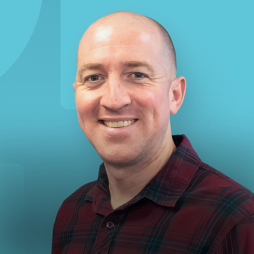 Smiling bald man in a red plaid shirt against a teal background.